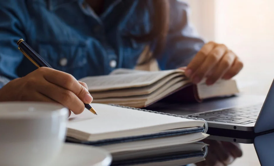 A woman writing in a notebook at a table with a laptop & coffee is engaged in writing and creating.