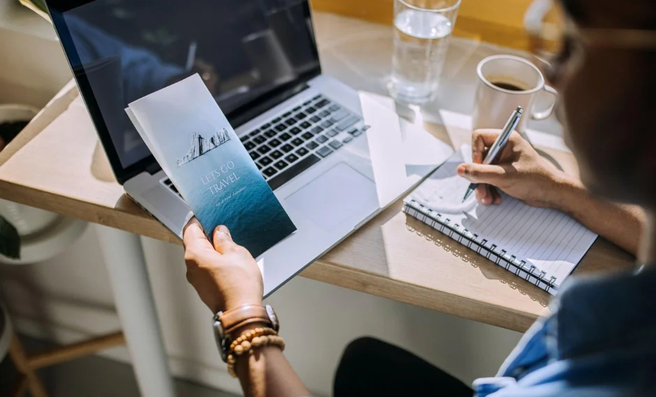 A woman writing content for a brochure while multitasking with a laptop, notebook, and pen.