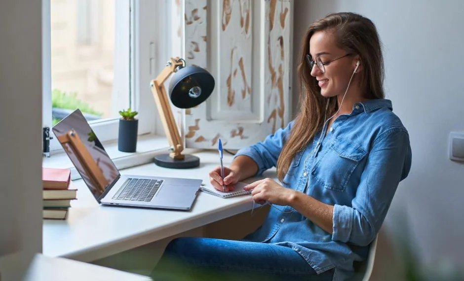 A woman sitting at a desk with a laptop and pen then she take notes down in a notepad what she hears